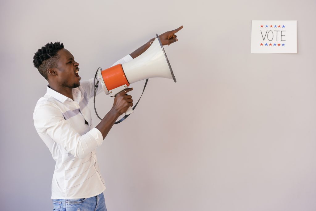Image Description: A Black man shouting through a megaphone and pointing at a sign that says vote. 