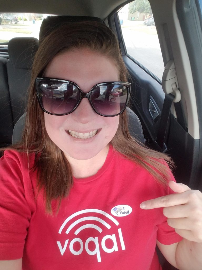 Image Description of a young woman with reddish hair and wearing sunglasses. She is sitting in a car wearing a red t-shirt with the white Voqal logo on it pointing at an I Vote sticker. 
