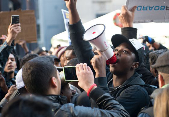 Image Description: Photo of crowd protesting during the day. The image is centered on a Black Man shouting upwards through a megaphone.