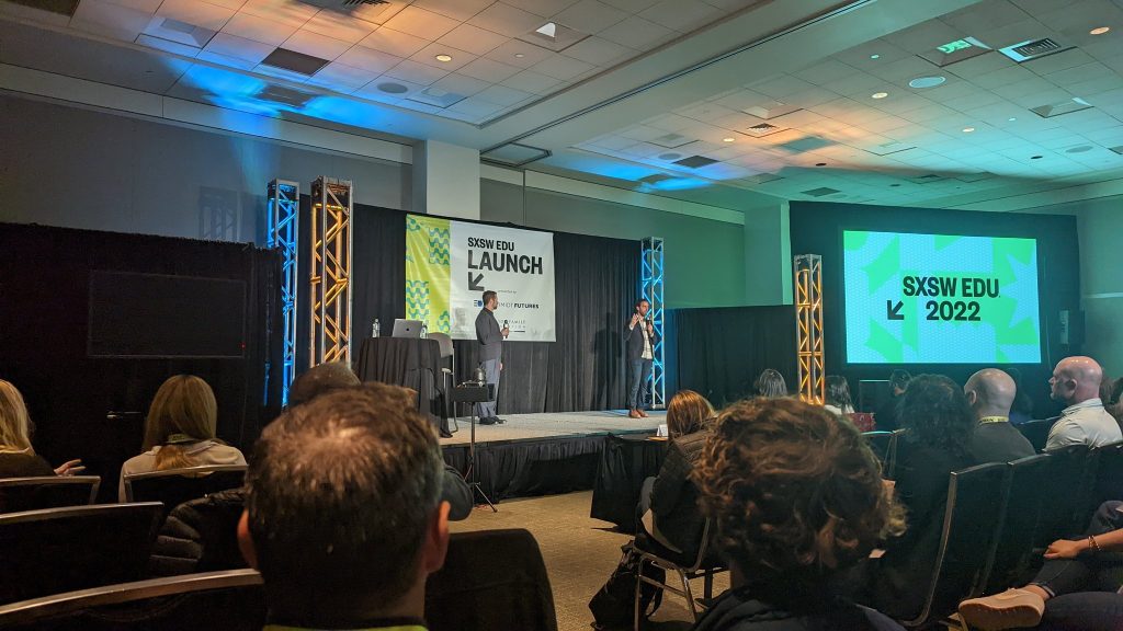 Image Description: SXSW EDU Launch Startup Competition Stage. Includes two individuals standing on the stage and a screen to the right. 